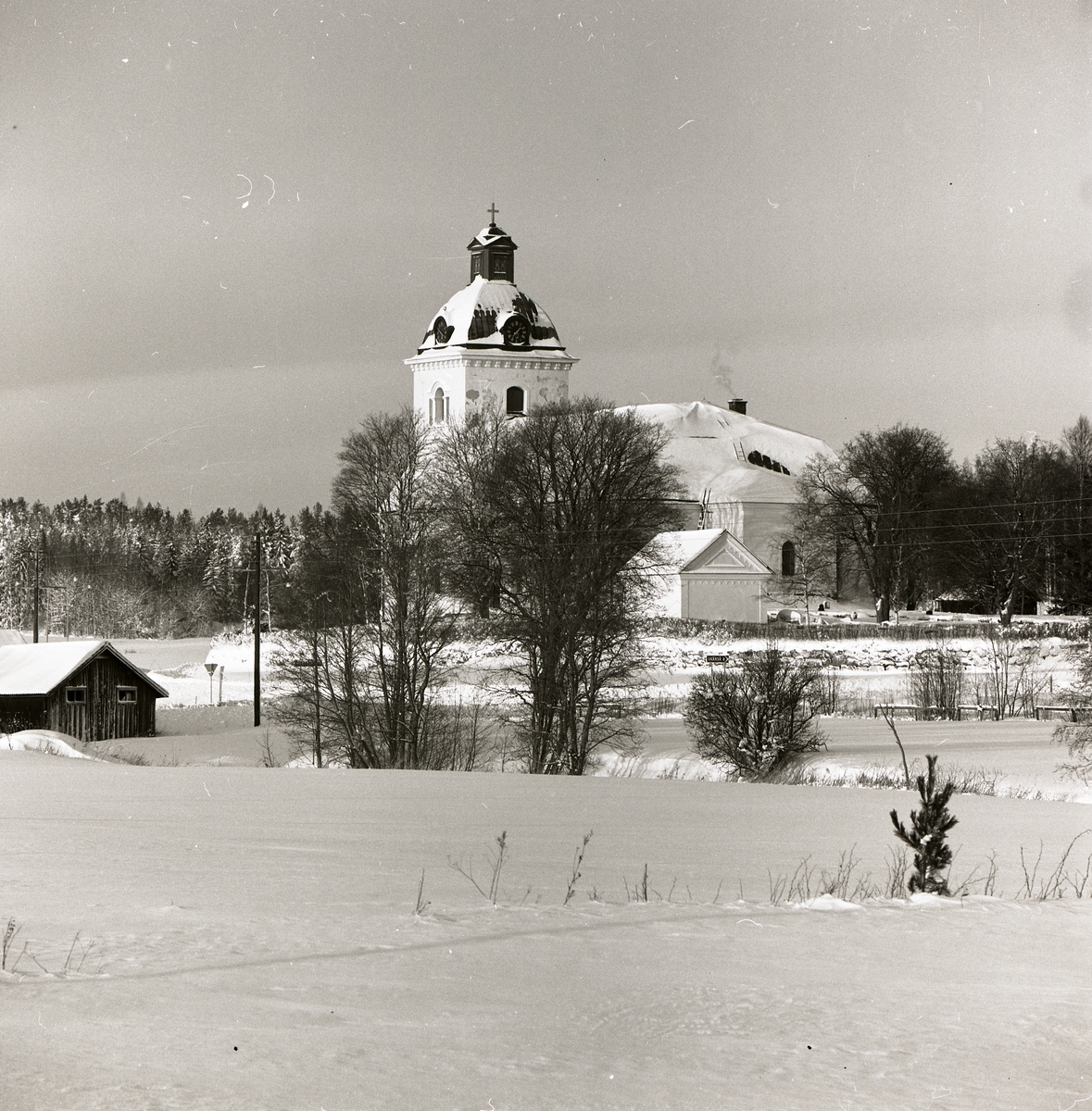 Norrala kyrka i vinterskrud syns bortom en åker varpå en liten lada står, 19 januari 1984.