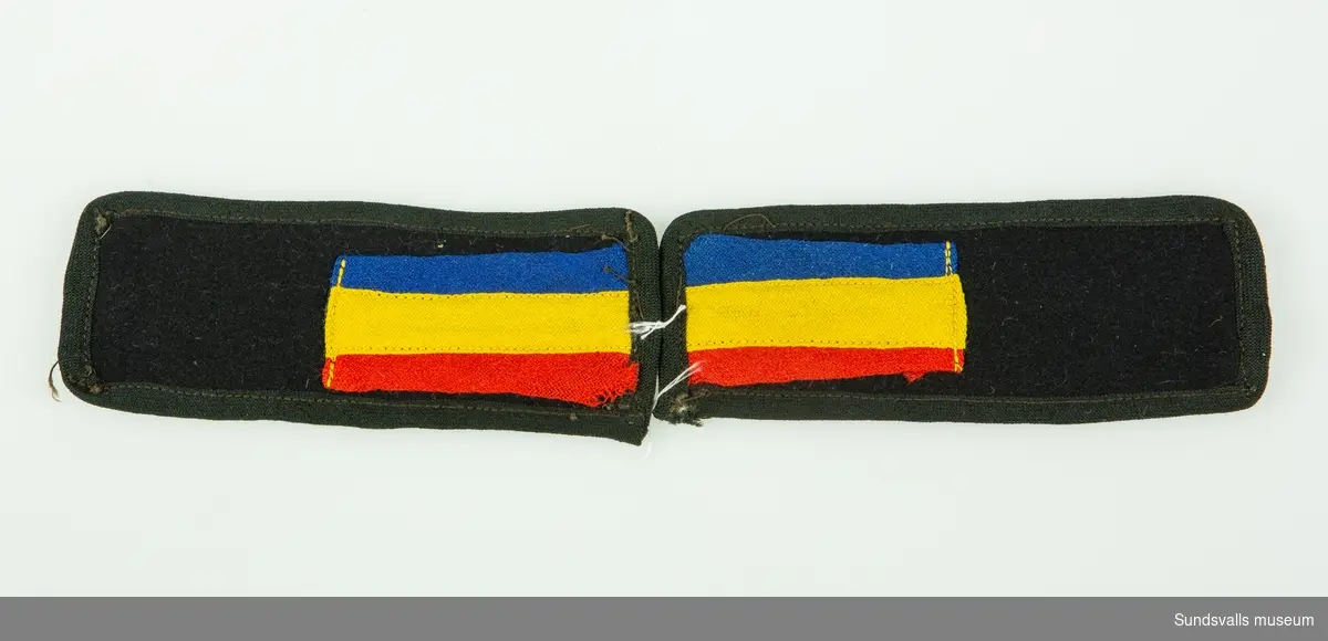 Axelklaffar av svart ylle med ett rött-, gult- och blårandigt band i ena änden på båda klaffarna.
