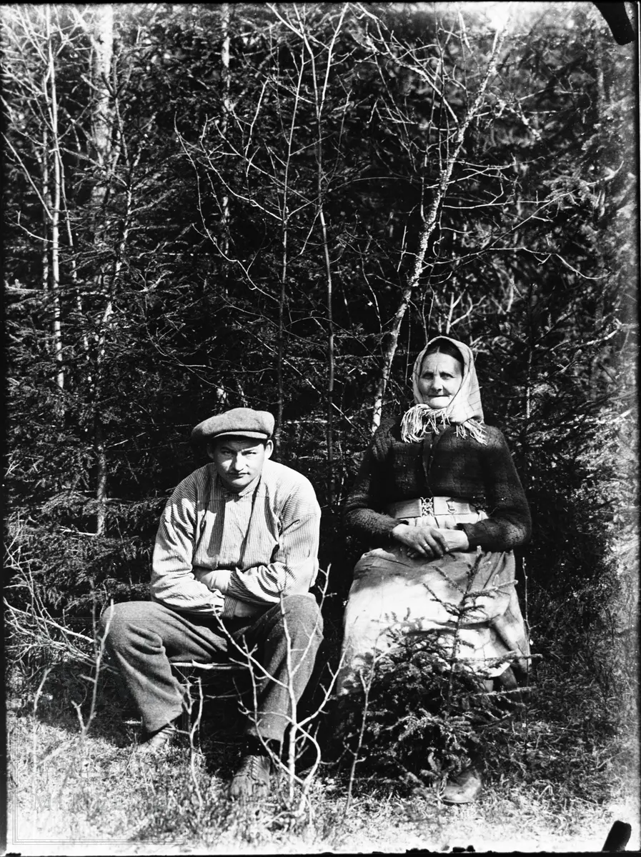 Eldre kone med skaut og en kar i buserull og sixpencelue sitter ute foran en skog.