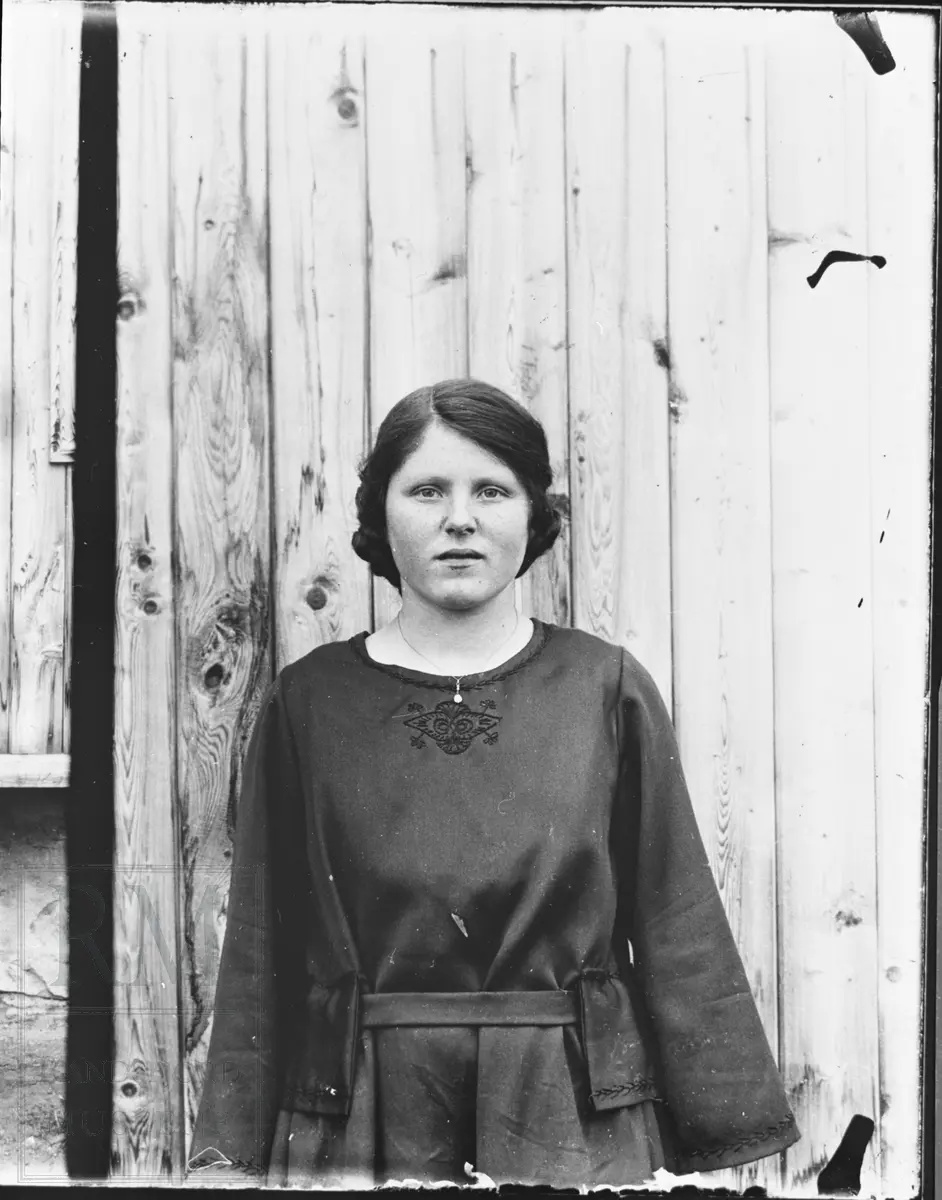 Portrett av ung kvinne, stående foran en husvegg, i mørk kjole.