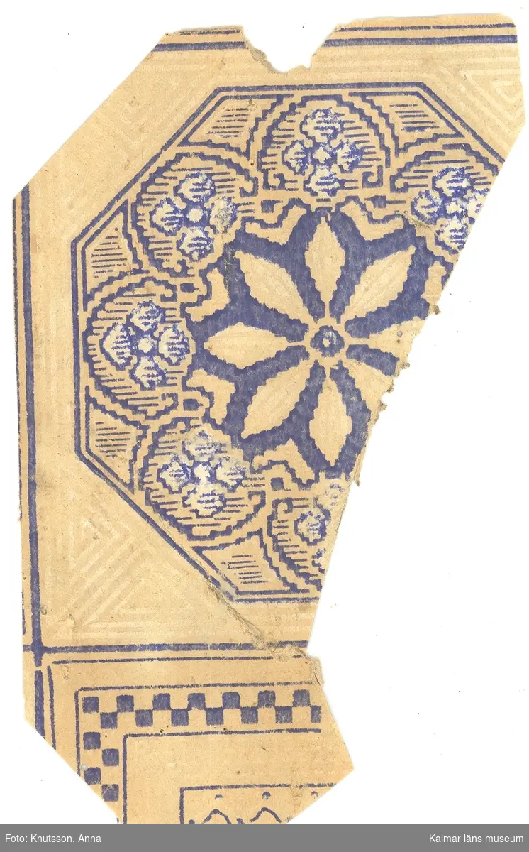 KLM 44026:1 Tapet i papper. Tapeten har pappersfärgad bakgrund och fyrkantiga inramningar i mörkblått. Eventuellt en kakeltapet. Mönstret i rutorna är stiliserade blommor i mörkblått och vitt. På baksidan: tidning med årtalet 1892.