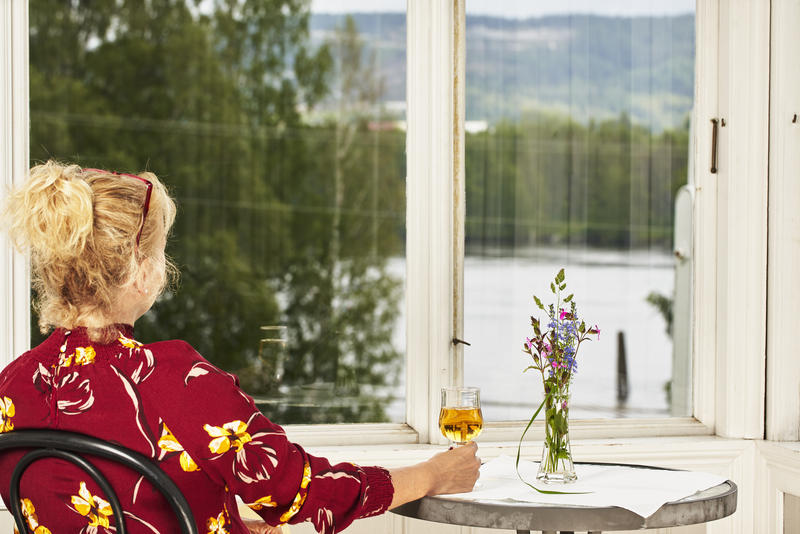 Dame i rød bluse som med ryggen til. Ser ut av vinduet ut mot elva.  Damen sitter på en stol og holder høyre hånd rundt et glass med eplemost som er plassert på et bord. En glassblomstervase med blomster (Foto/Photo)