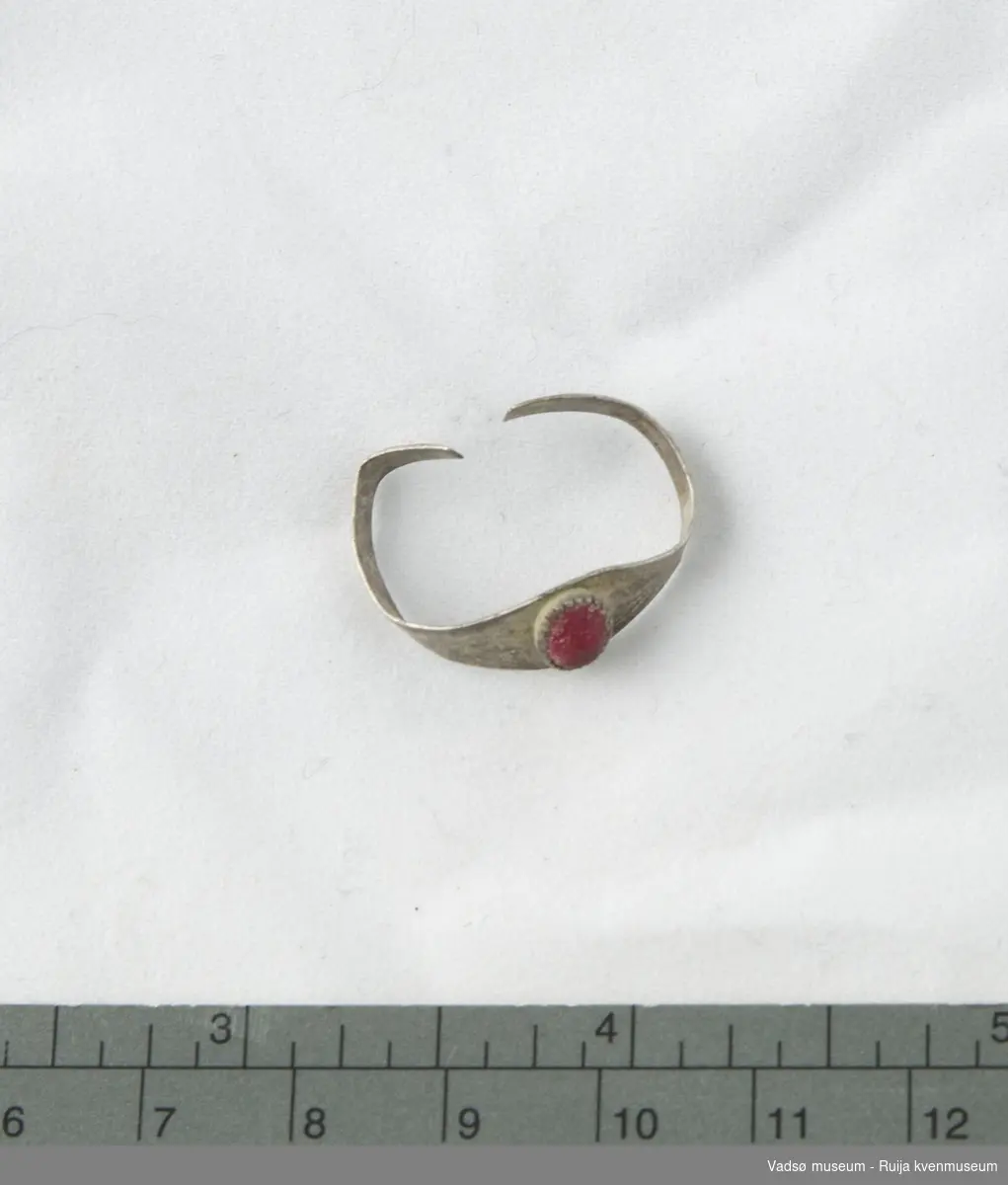 Sølvring som er dekorert med en oval, rød smykkestein av glass. Steinen har avrundete kanter, men viser tegn til opprinnelig fasettering. Sisellert prikke- og strekdekor på ringen.