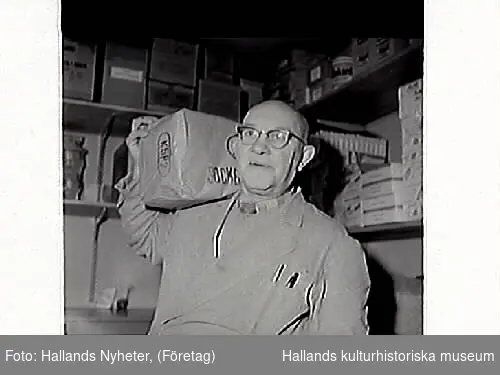 John Johansson står vid en trave ägg i Konsum Frillesås. I bakgrunden står rullar med vad som kan vara prover på olika linoleummattor.
Bild 2 visar John med en i papp inslagen sockerpaket på axeln.
