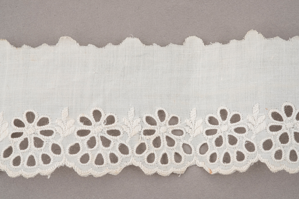 Hvit tekstilbånd med hullbroderinger av blomster langs den ene kanten. Den andre kanten er klipt opp i bølger. Endene er rettklipt.