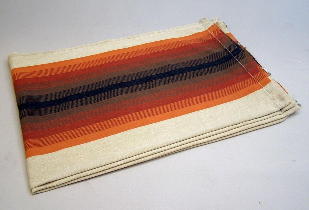 En gardinluft bestående av två stycken längder, bomullstyg med mönster av ränder av olika bredd i färgerna: beige, orange, röd, rödbrun, brun och svart. I ovankant är metallhängare fastsydda för upphängning.