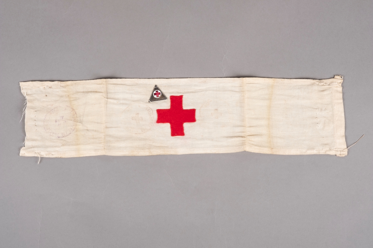 Et armbind i hvit bomull med påsydd rødt kors i tekstil. Ved korset er det påsydd et hjelpekors-merke. Det er stempler på armbindet som er noe falmet.