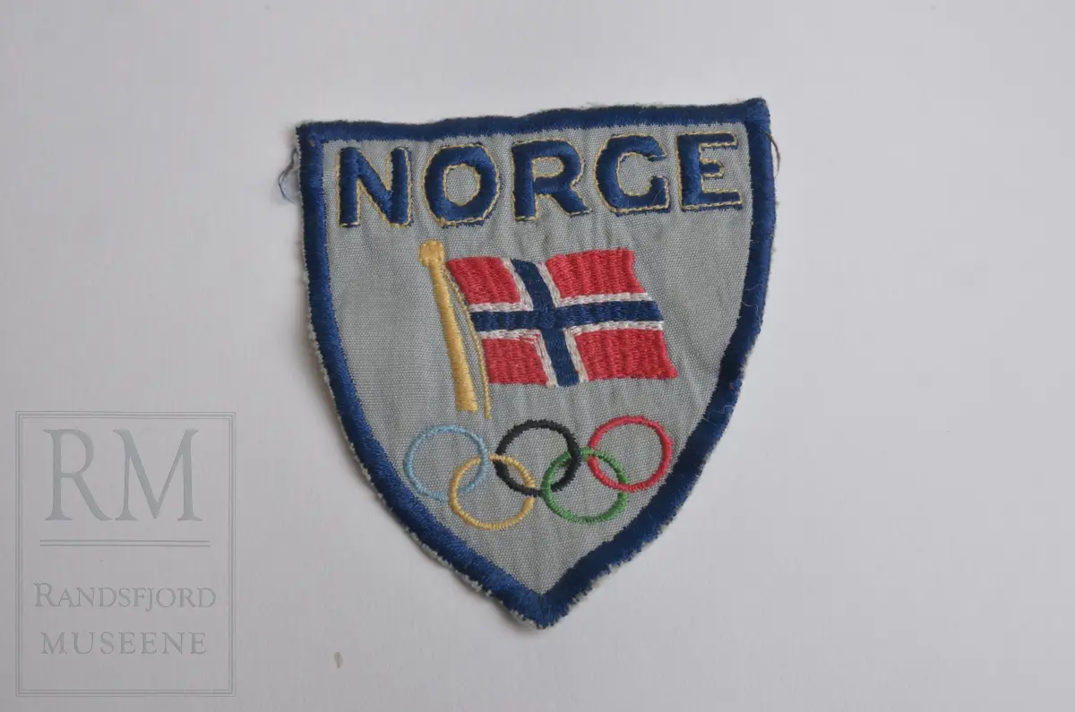 Grått stoff fôret med hvitt stoff bak. Brodert blå kant, NORGE, OL-ringene og norsk flagg. Maskinsøm.