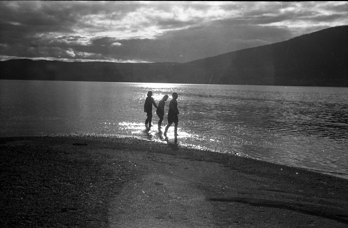 Avfotografert bilde av tre personer på en strand ved en innsjø. Hverken personer eller sted er identifisert.
