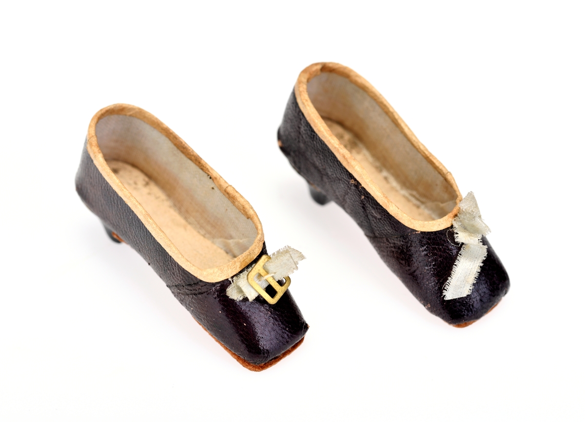 Et par dukkesko med høy hæl og firkantet tå. Skoen og hælen er laget av brunt papir som ligner på skinn (bokbindingspapir?). På undersiden er det limt på en lærsåle. Langs kanten oppe er det limt på en hvit papirkant. De har hvitt for og pappsåle. Foran på skoene er det festet hvite silkesløyfer. På den ene skoen er det også en gullfarget metallspenne.