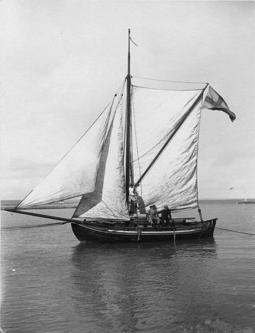 von Dübens segelbåt Blenda med fulla segel ligger förtöjd i Gränna hamn. Två män sitter i båten och en pojk hänger i en tamp.