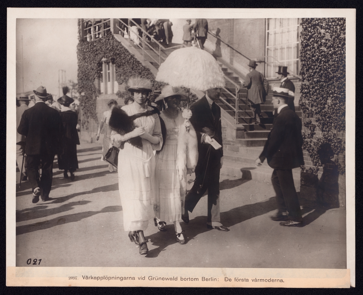Bilden visar finklädda damer och herrar som promenerar. I bildens centrum går två damer i vita klänningar med ett paraply som solskydd.