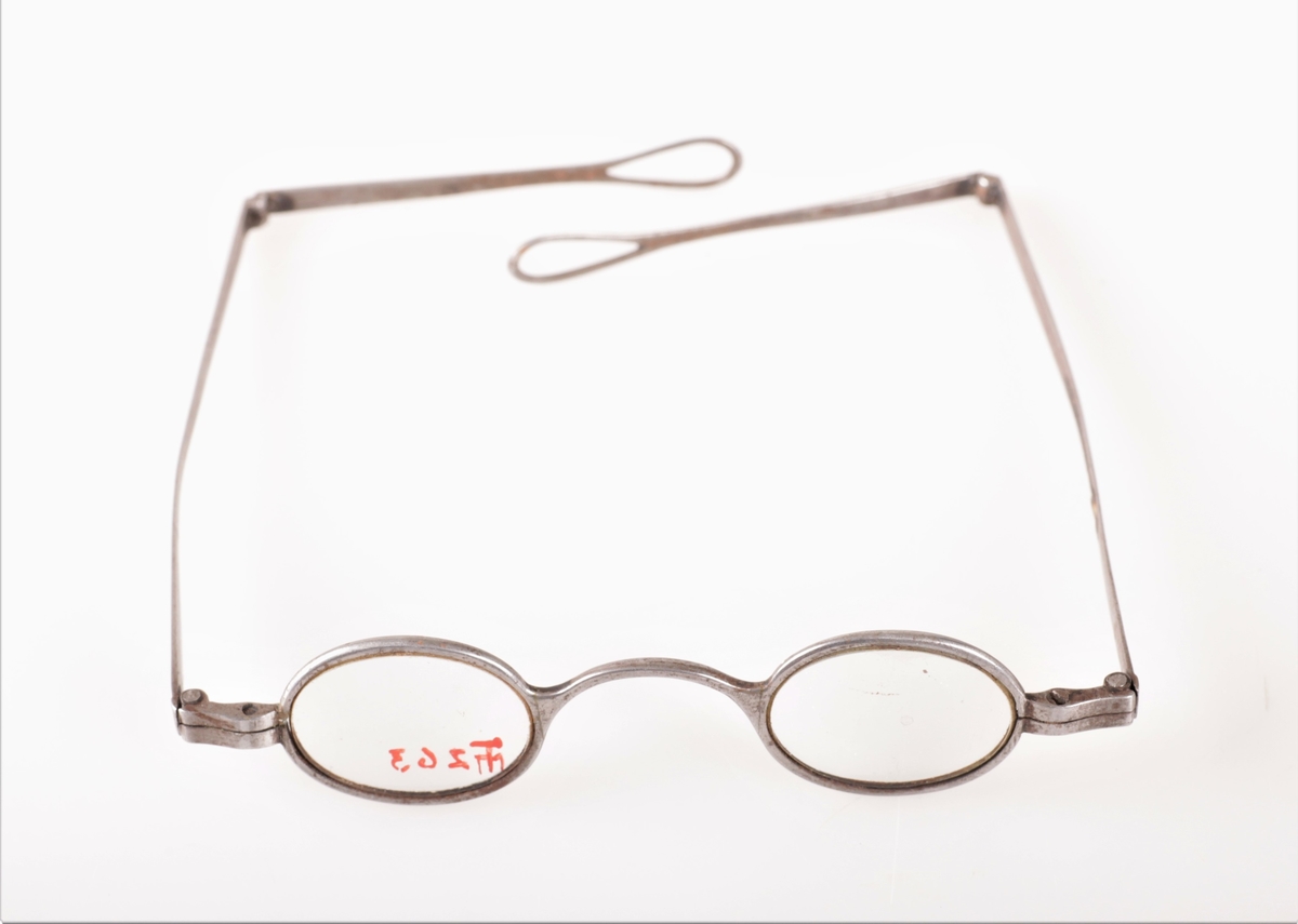 Ovale brilleglass i stålinnfatning, flate stenger med ledd som bøyer inn. Ytterst på leddet stort pæreformet hull