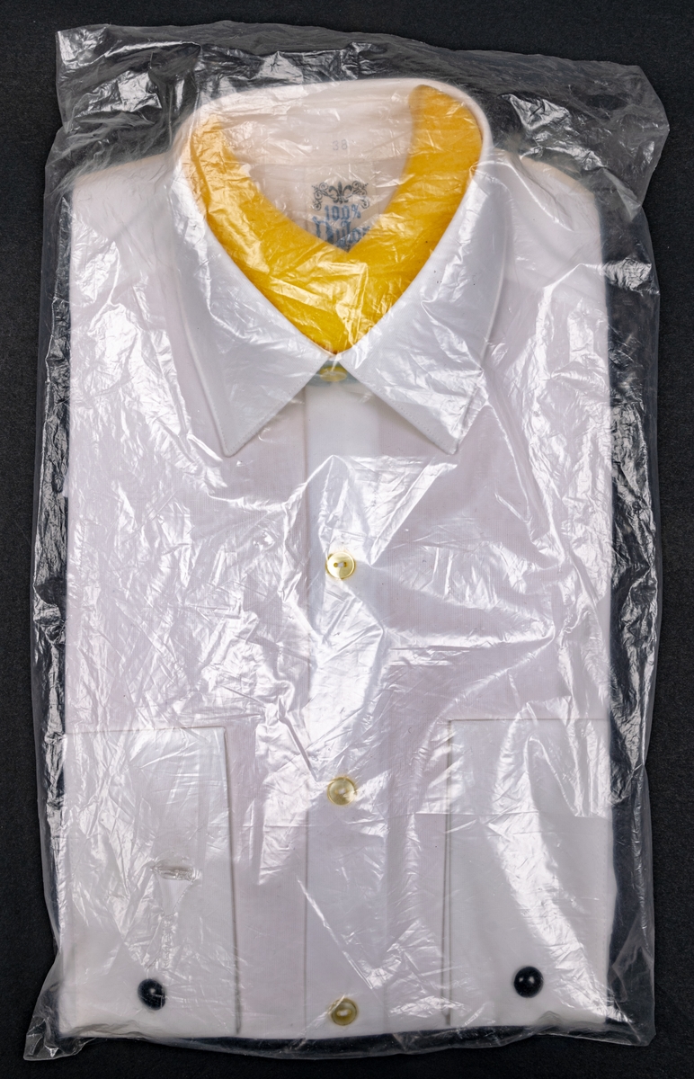Vit nylonskjorta i originalförpackning. Skumgummibit innanför halslinningen. Vita knappar och svarta manschettknappar.
