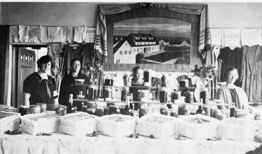 Elevutstilling ved Sømme Husmorskule i 1926. Framme ei rad med småkakeboksar, i bakgrunnen eit drapert maleri av skulen.