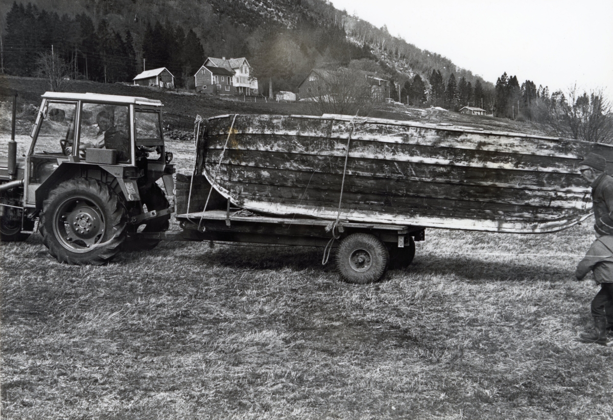 Motiv av en traktor som frakter en båt. Båten er en seksring som Sogelaget ved Dalsfjorden skule har engasjert seg i å bevare. I bakgrunnen er et gårdsbruk.