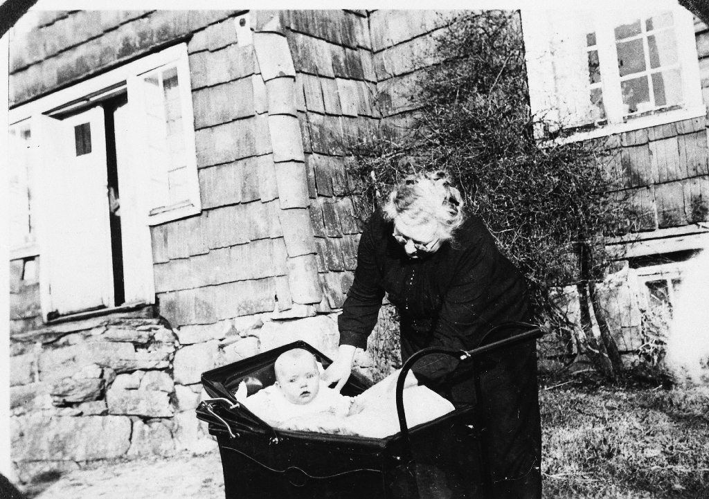 Grete Aasland med barnebarnet Ingfrid Håland i vogna framfor det pannekledde huset på Lindland.