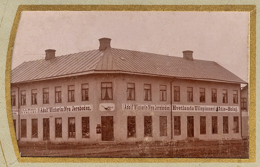 En större affärsfastighet i Vetlanda. På skyltarna står: "C.A. Winell, Färgare", "Adolf Wictorin: Nya Jernboden", "Hvetlanda Ullspinneri Aktie-Bolag".