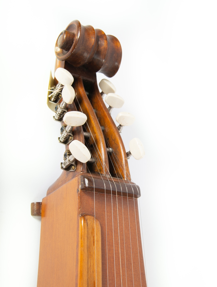 Langeleik i eske. Instrumentet er svakt svunget og dekorert med utskårne hjerteformer i treverket/ lydkassa. Halsen svunet og med hulkiler. Løse stemmeskruer i tre og streng ligger i liten plastpose