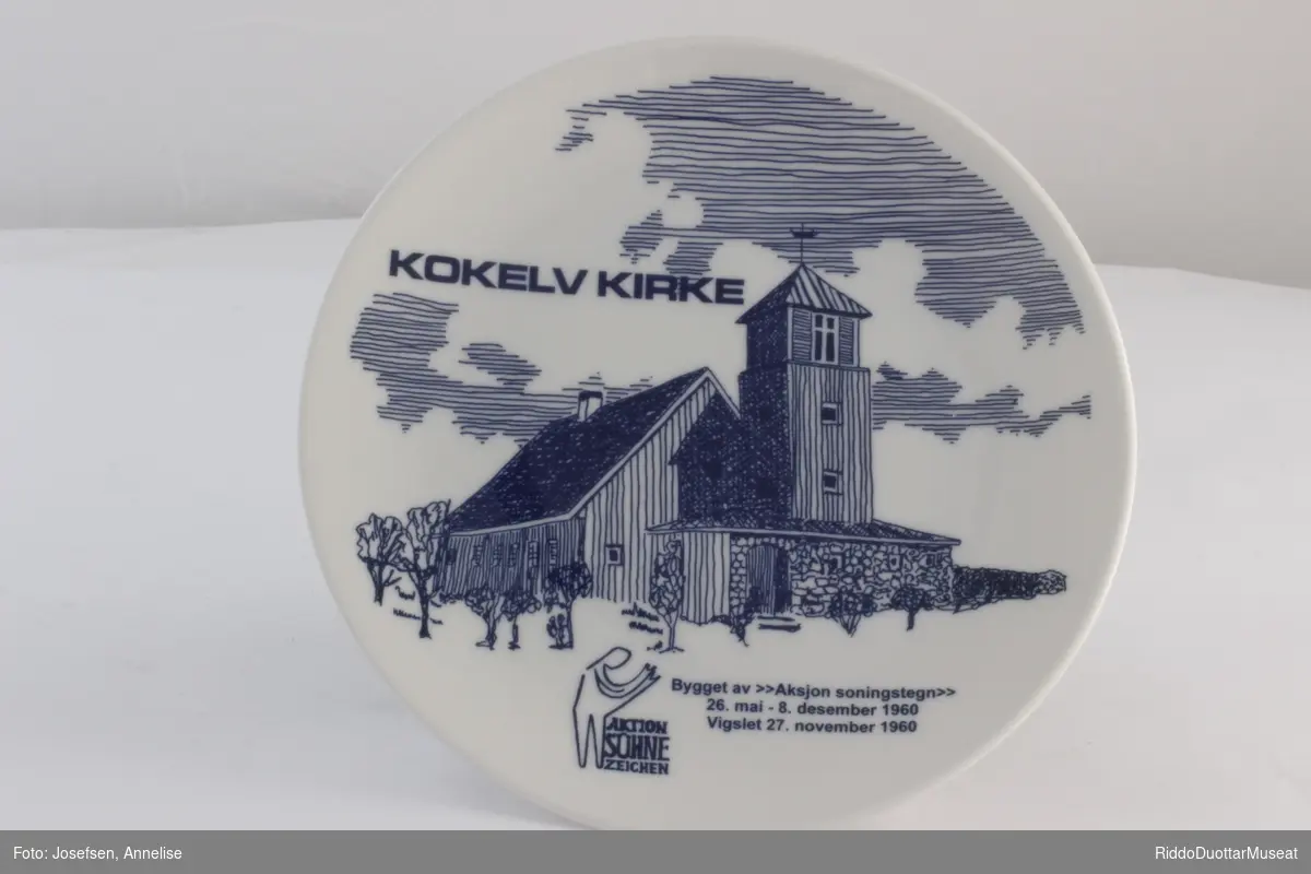 Hvit porselensfat med tegning av Kokelv Kirke. Blått motiv og logo for Aktion Sûhne Zeichen. Oppheng bak og tekst med alle som deltok i byggingen på baksiden.