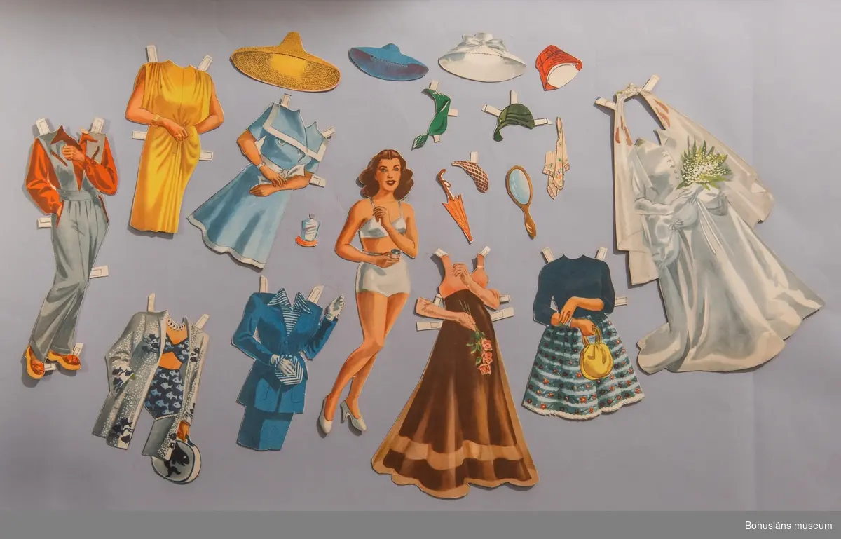 Pappersdocka i form av vuxen kvinna med tillhörande åtta klädesplagg varav en brudklänning, en aftonklänning, en sportdress, en stranddress, en dräkt, en eftermiddagsklänning, en vardagsdress med kjol och jumper (sitter ihop), en vardagsklänning, en cocktailklänning i gult. Åtta hattar för fritid, vardag och fest, ett paraply, en handspegel och en parfymflaska.
Förvaras med UM026095