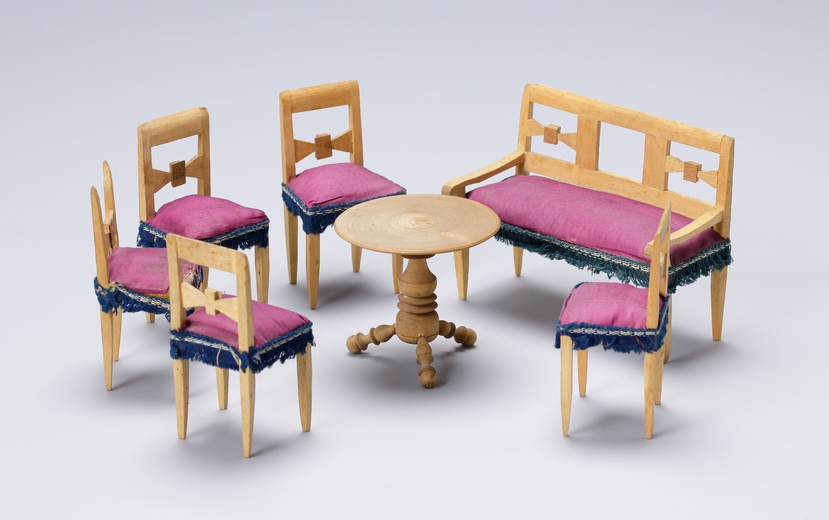 Dockskåpsmöblemang av furuträ bestående av fem stolar, en soffa och ett bord, klädda med lila siden. Runt kanterna band och frans av blått bomullsgarn. Bordet svarvat.

Inskrivet i huvudkatalog 1938.