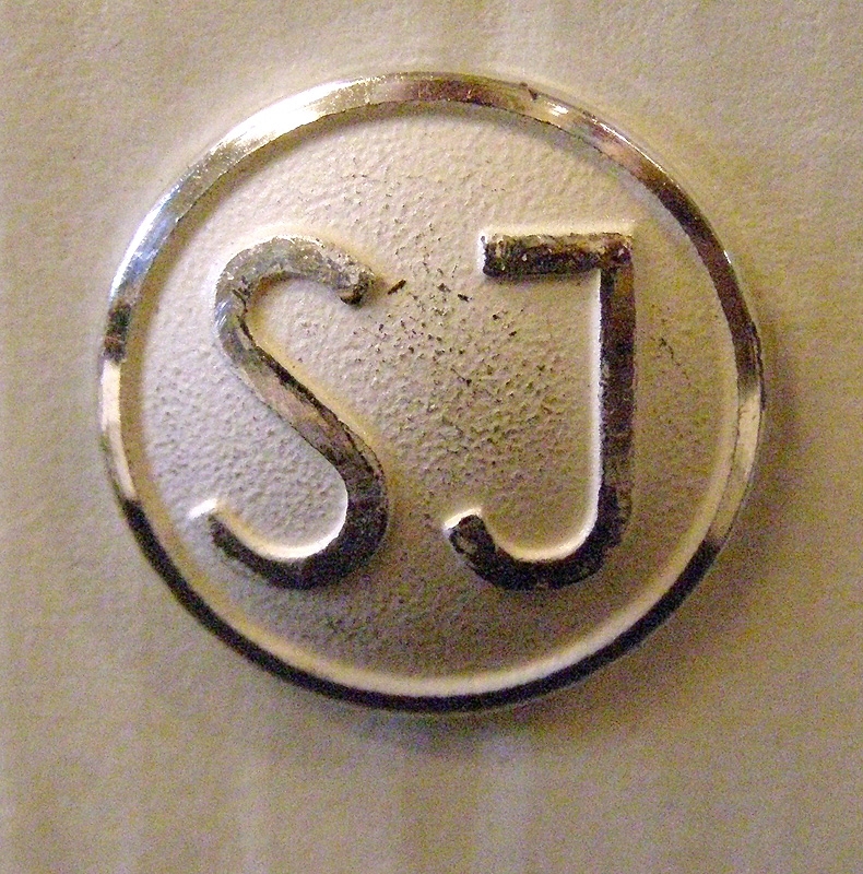 Två mösstränsknappar av silverfärgad metall, med SJ:s logga i form av initialer. Knapparna är något större än vad de egentligen ska vara.