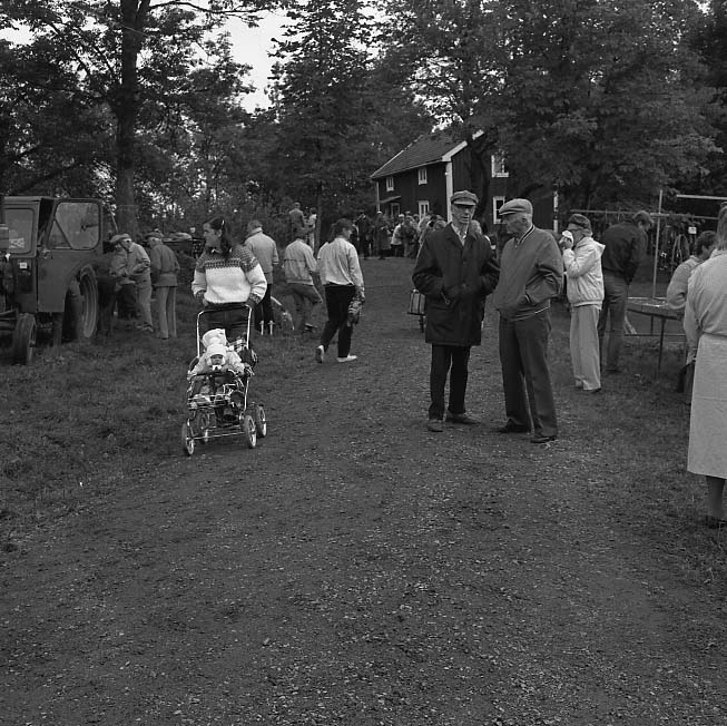 Auktion på okänd plats. Människor kantar en grusväg upp mot en bostad. En ung kvinna med barn i vagn promenerar mot fotografen.