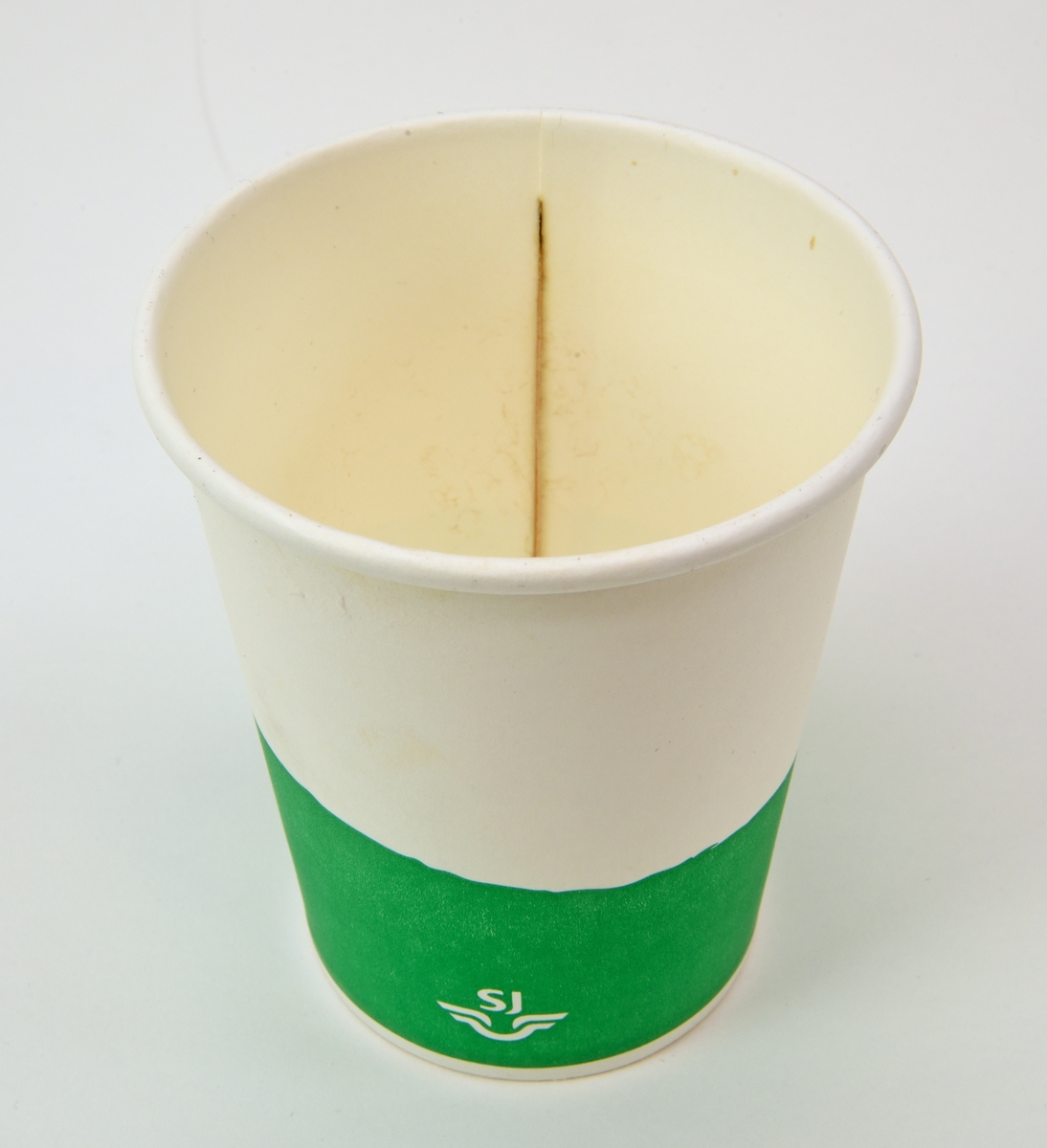 En pappmugg med lock av plast från SJ Bistro. Muggen har ett grönt fält nedtill med vit SJ-logga. Locket är vitt med ett avlångt hål. Muggen har spår efter te inuti.
