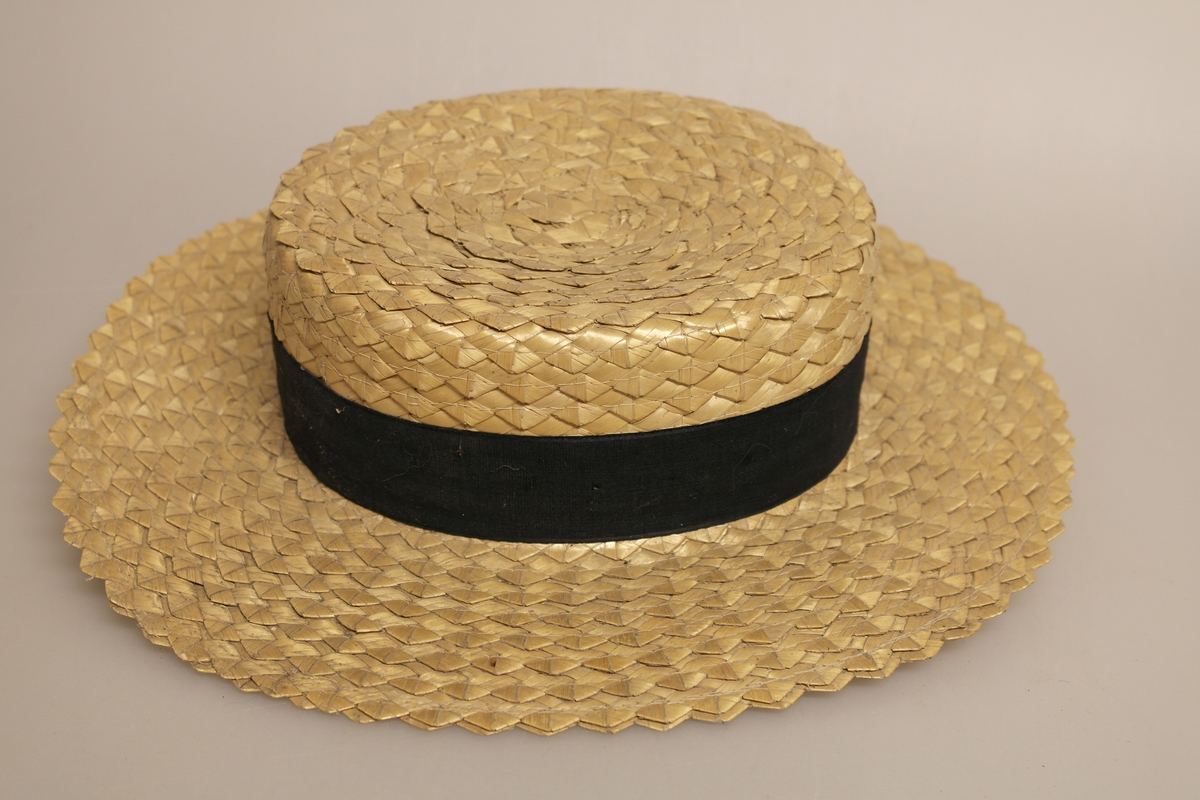 Naturfarget, oval stråhatt med flat pull. Hatten er sydd av flettede bånd av halm og har svart ripsbånd med sløyfe, samt hvit svetterem.