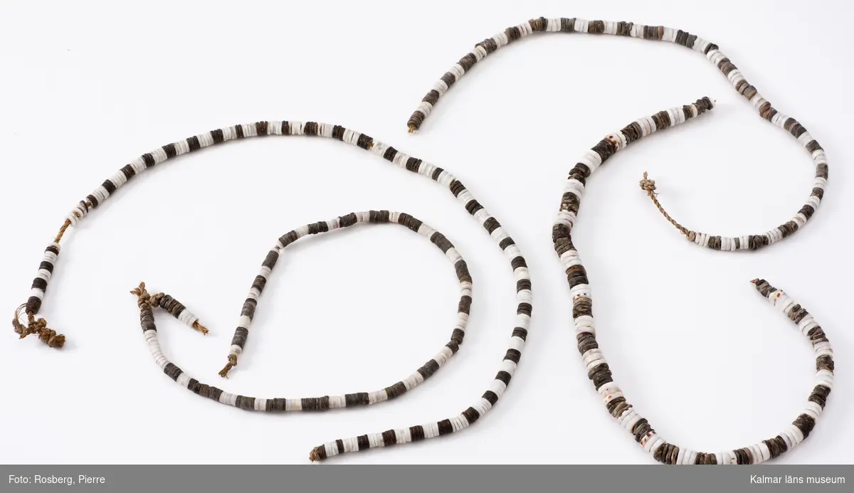 KLM 4046. Pärlor av snäckskal eller kokosnöt. Pärlorna uppträdda på fyra stycken snören. Pärlornas diametern växlar mellan 0,5-1 cm, uppträdda på fyra snören med längd 47 cm, 40 cm, 38 cm respekive 38 cm. Vita och svarta ringformiga pärlor, samlade på fyra snören av bastfibrer, de vita är av snäckskal, de svarta av kokosnötskal.