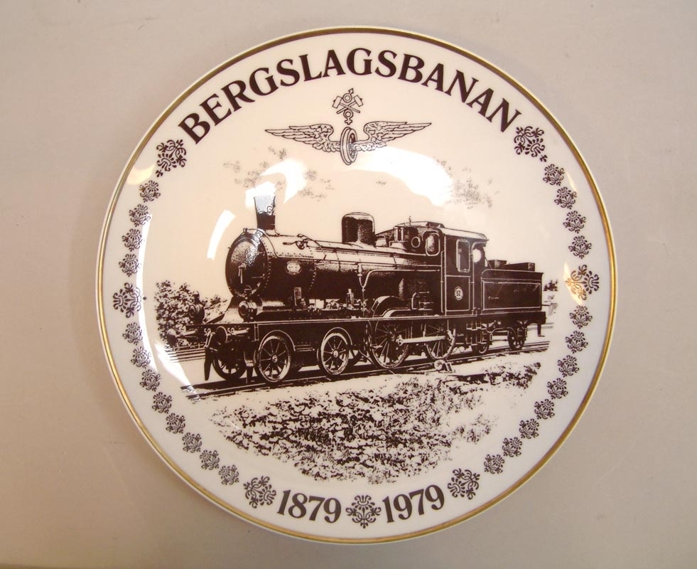 Vit porslinstallrik med brun dekor och förgylld kant. Text: "BERGSLAGSBANAN 1879 - 1979"

"Hjubileums Hjultallrik"
Nummer 185 av 300.

Bilden föreställer BJs lok C3 NR 52.