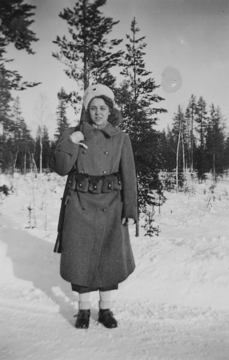 Porträtt av luftbevakare Inger (efternamn okänt) i uniform och med gevär, vintern 1942. Utomhus vid luftbevakningsförläggningen för 91:a ls-kompaniet i Tellejåkk, Kåbdalis under beredskapsåren.