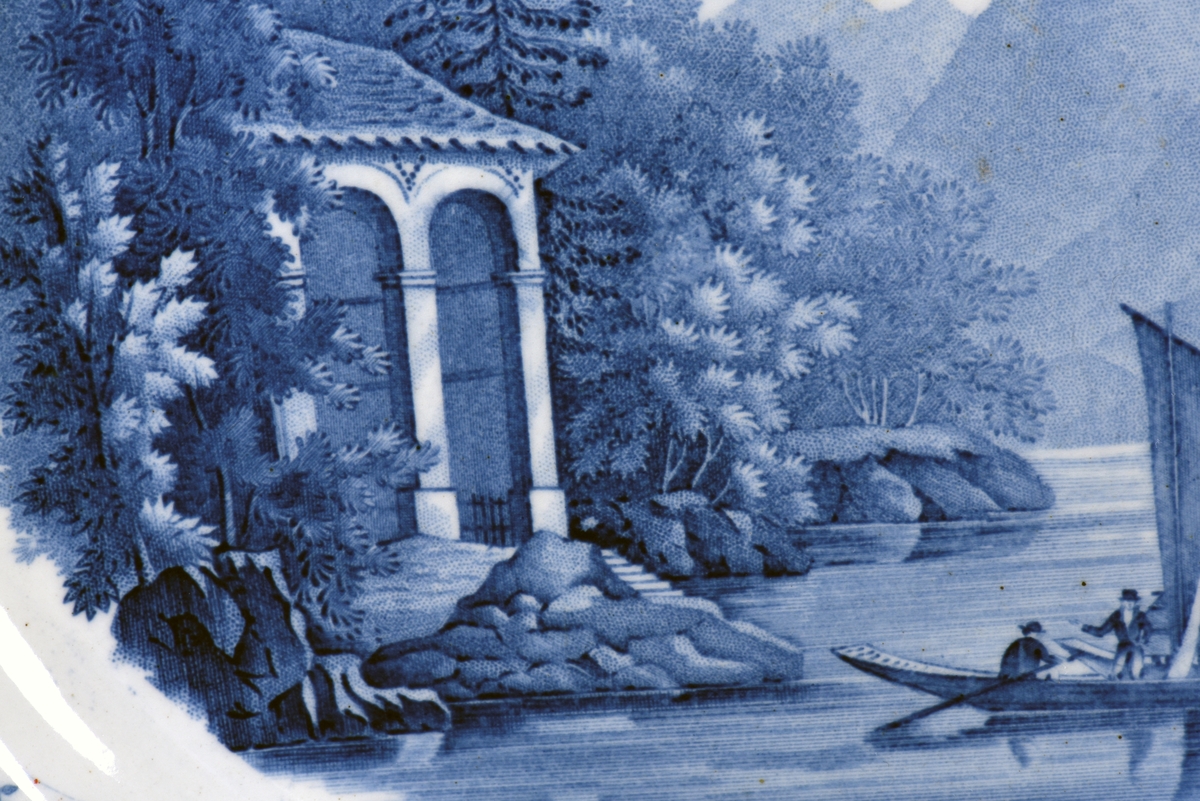 En av sex flata tallrikar tillverkade av Rörstrand. Vit tallrik med blå dekor med ett alplandskap i bakgrunden och en sjö med segelbåt och ett tempel i förgrunden. Runt brämet dekorerad med en blomsterrranka med bland annat rosor.