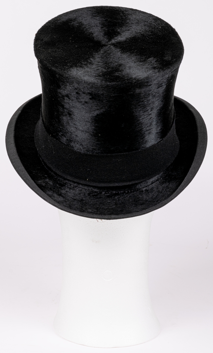 Stormhatt märkt Aktiebolaget J.W Haglund, N. Kungsgatan 1, Gefle. Svart.
Höjd 14cm, brättbredd 4,5cm.
Hattask av svart papp med guldtext på locket. English manufacture of hats. Best London deposed.
