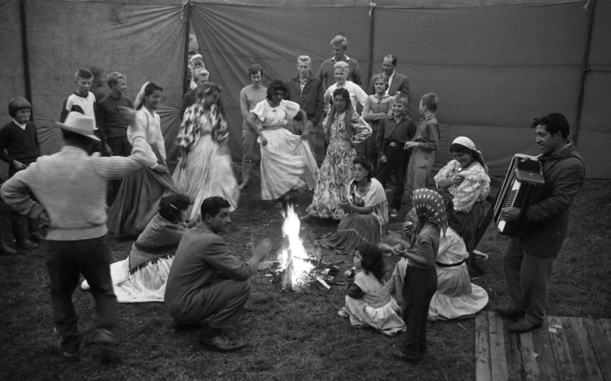 En grupp romer dansar och spelar musik kring en brasa. I bakgrunden öppnas en tältduk och en grupp människor kommer in.