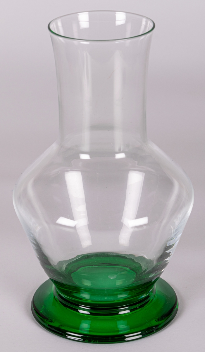Vas av ofärgat glas på låg, utkragad fot i grönt glas. Delvis konisk med hög, smal hals. Design Gunnar Cyrén för Orrefors Glasbruk.