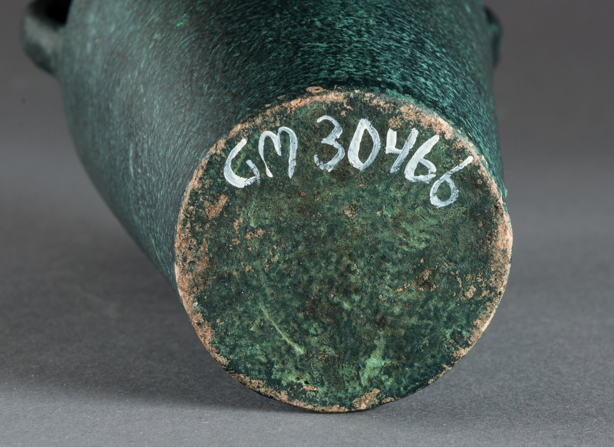 Vas med två hänklar av lergods. Handdrejad och glaserad och målad i s.k. grön patinering.
Vasen är omärkt men förmodligen från Bo Fajans i Gävle.