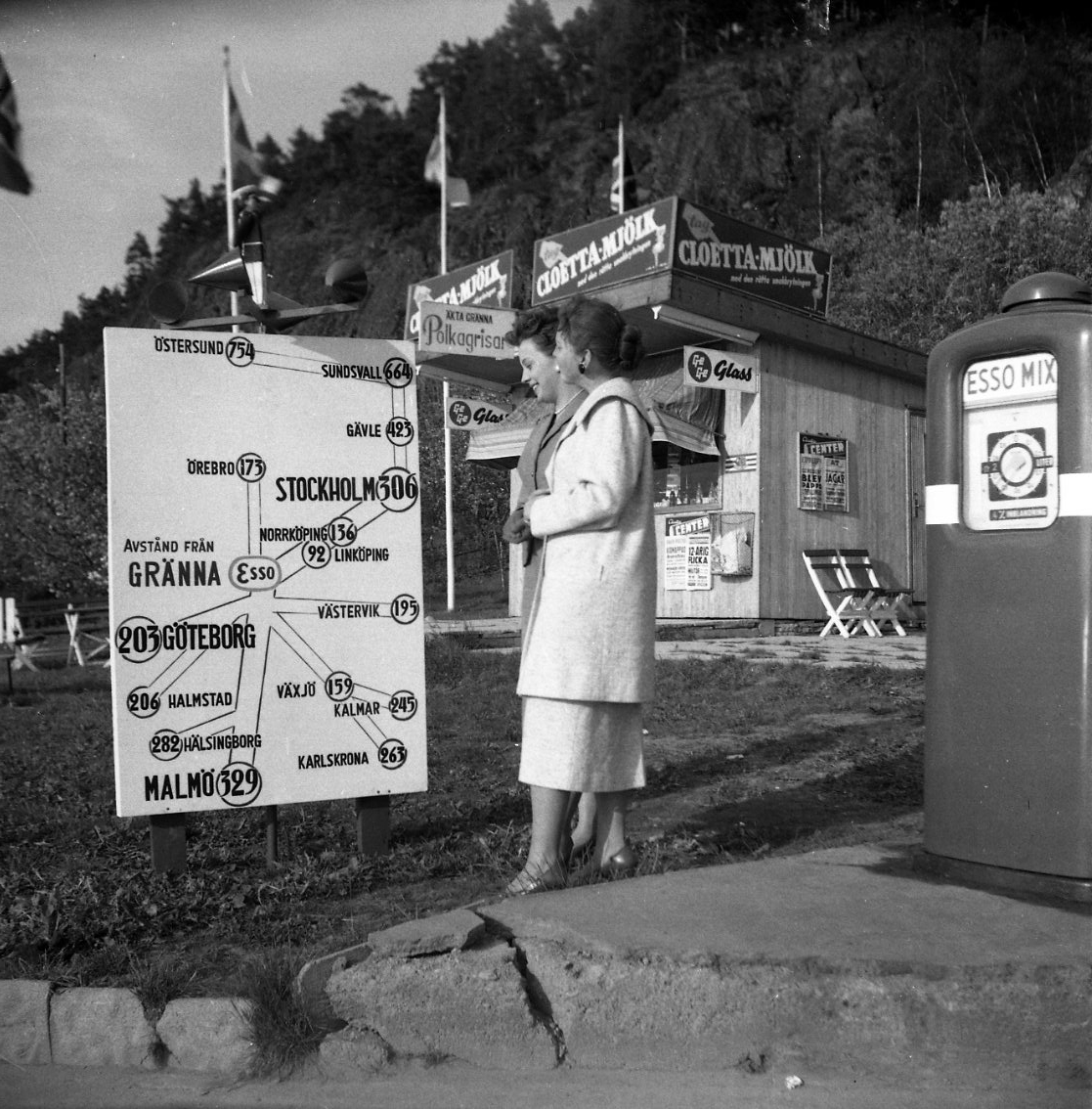 Två kvinnor i kappor står och tittar på en vägskylt vid Esso-stationen i Gränna som visar vägar och avstånd till bland annat Örebro och Stockholm. Bakom dem en liten kioskbyggnad med reklam och skyltar för "Ge Ge Glass", "Cloetta-Mjölk" och "Äkta Gränna Polkarisar". Till höger en bensinpump "Esso Mix.