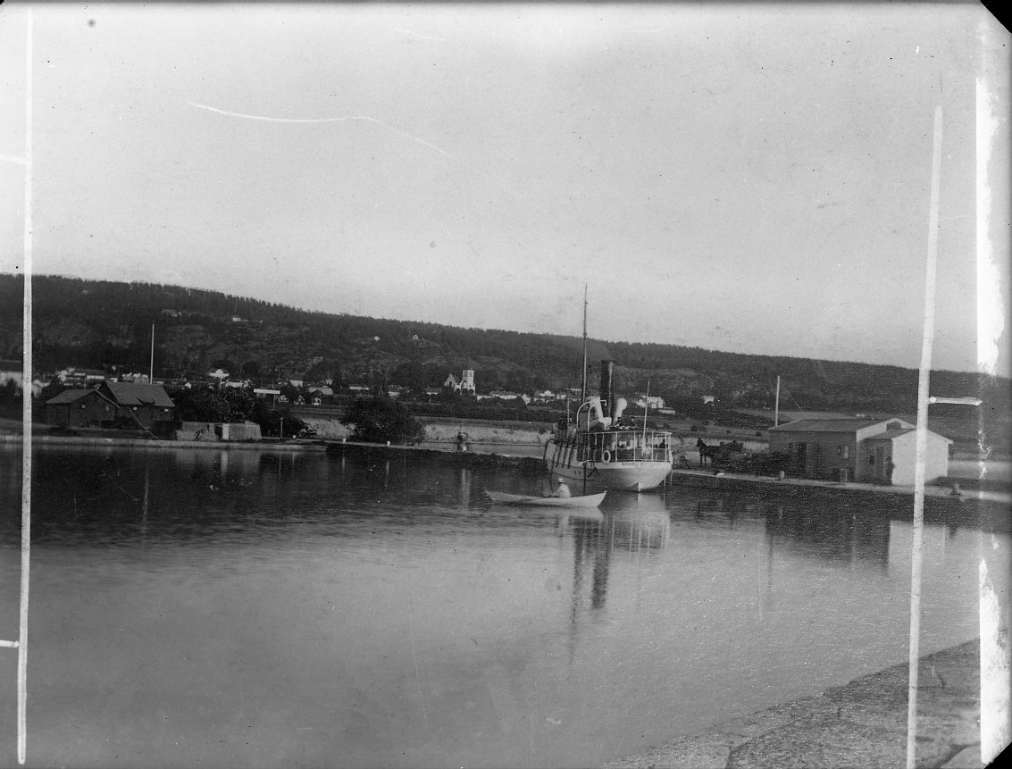 En ångbåt ligger vid kaj i Gränna hamn. En roddbåt passerar. Ångbåten är identifierad som Södra Wettern som gick regelbunden trafik mellan Gränna och Visingsö.