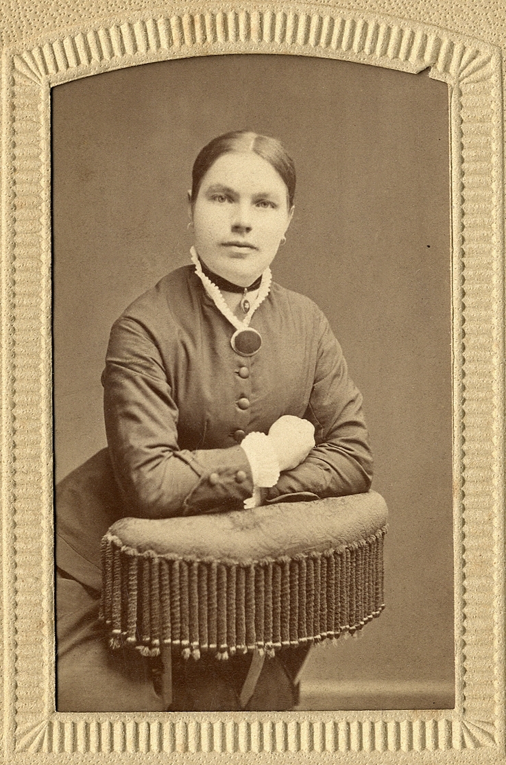 En okänd kvinna i mörk klänning med vit kråskrage, lutar sig mot ryggstödet på en stoppad stol med frans.
Knäbild, halvprofil. Ateljéfoto.
