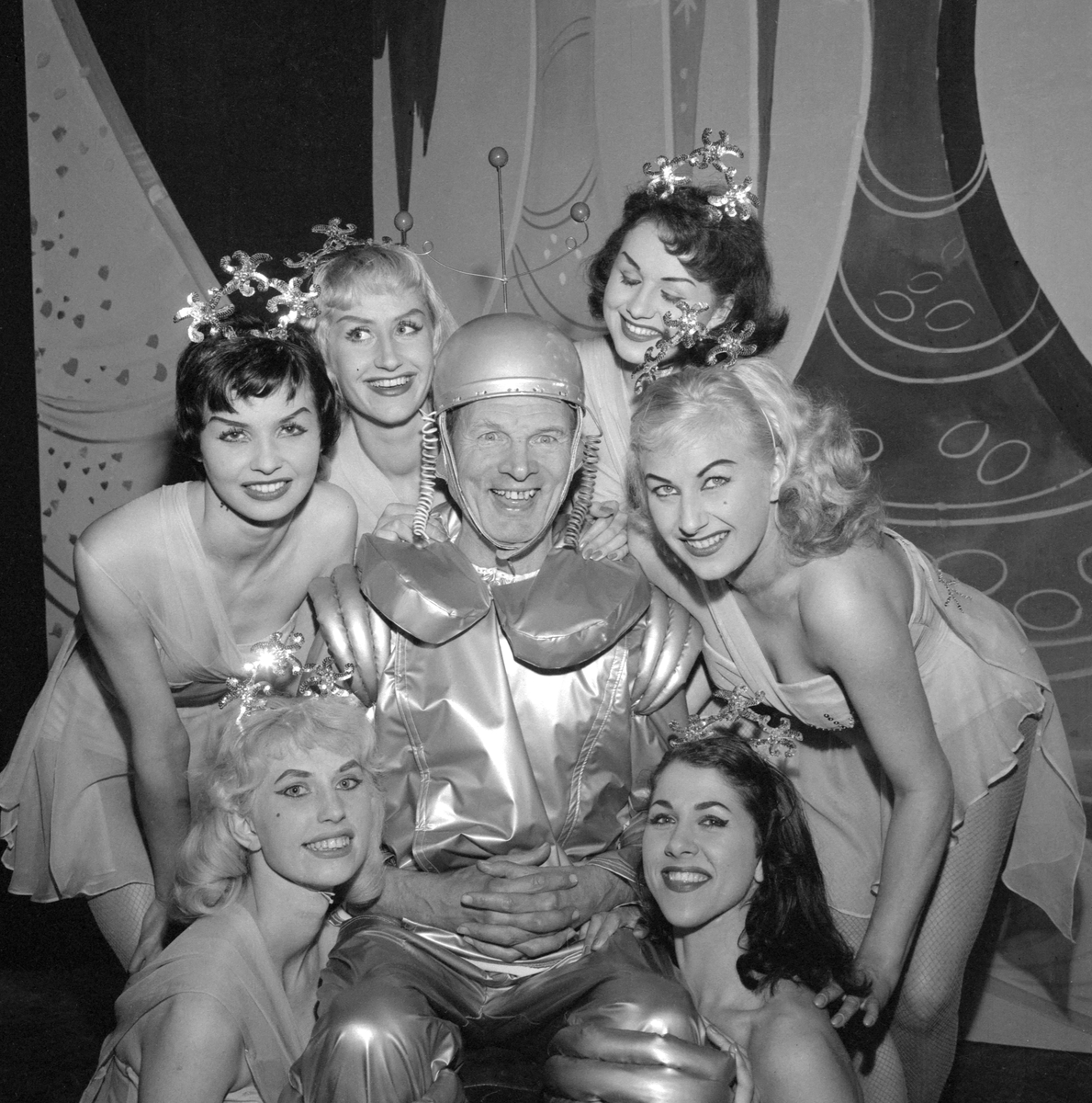Ett besynnerligt sceneri som har sin förklaring i 1959 års Klangerevy. Gustav Lövås är skrudad för revynumret "Träff på Paradisplaneten" och omkring sig har han samlat ensemblens dansare, medlemmarna i den internationella balett-truppen Roxy Girls. Bilden är från revyns premiär i Mjölby folkpark.