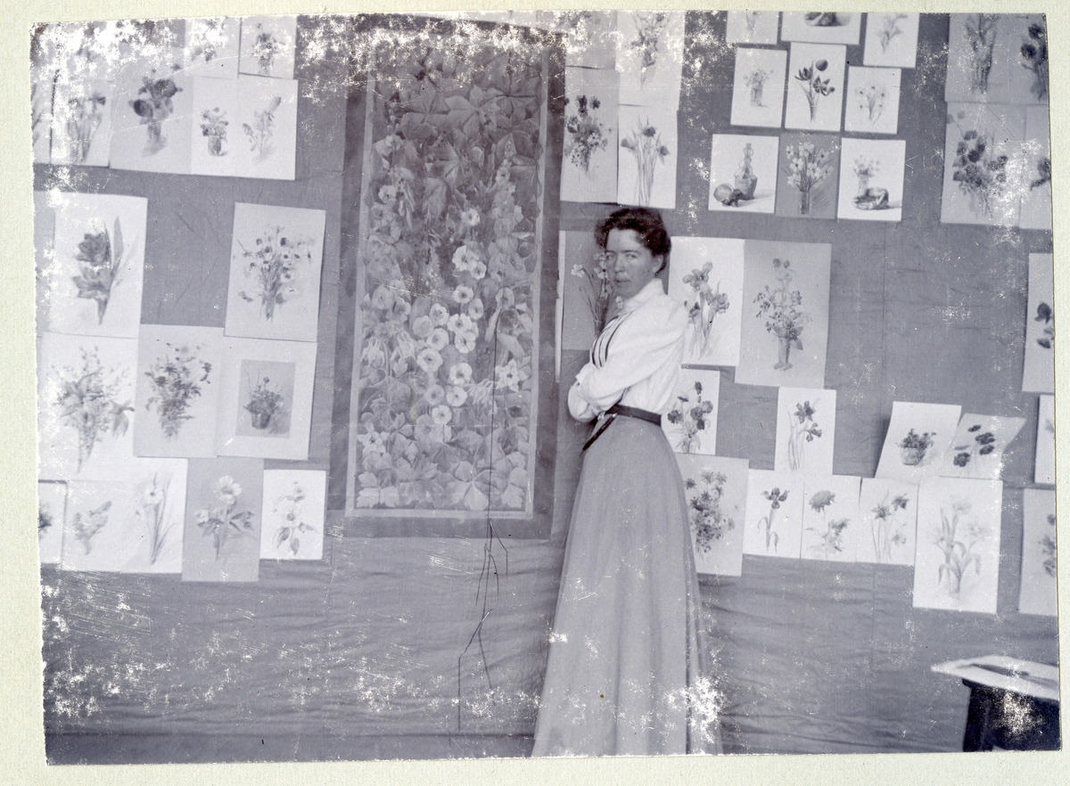 Stockholm. 
Kvinna stående framför vägg med blomstermålningar. 1901.