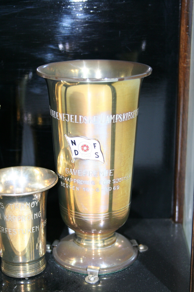 Pokal med innskrift og logo. Pokalen står skrudd fast til bunnen av monteren med tre klemmer.