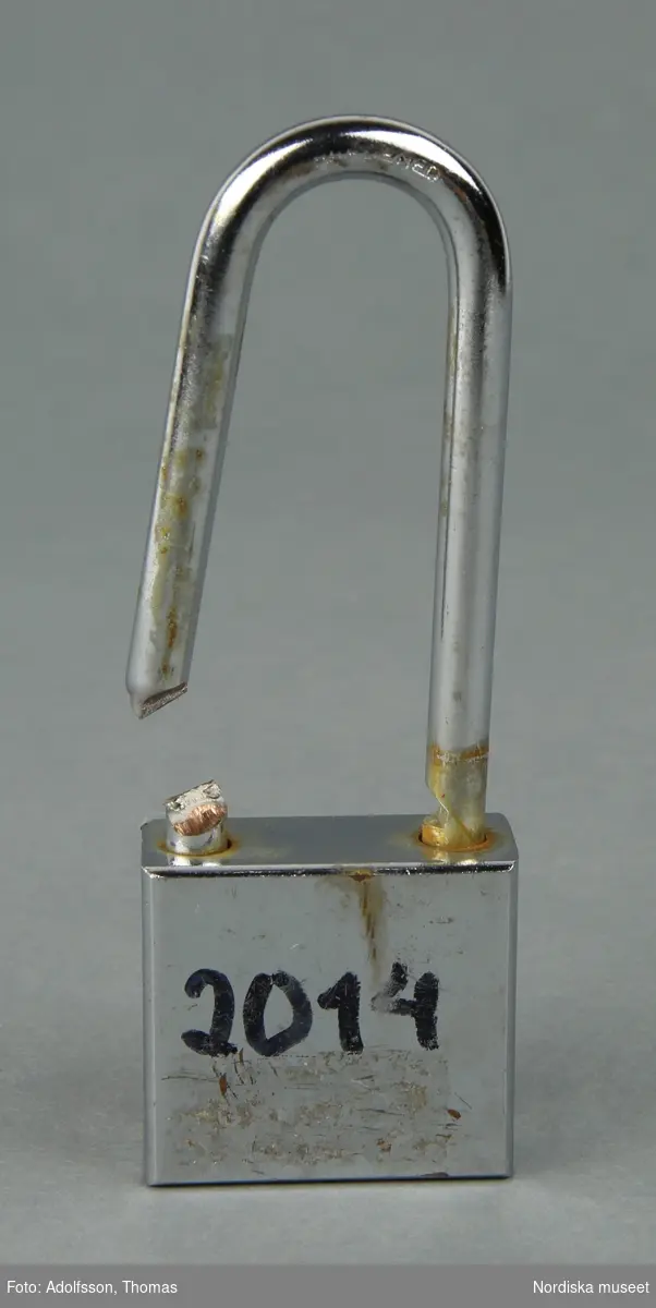 Ett fyrkantigt avknipsat silverfärgat kärlekslås av metall, med namn, årtal m.m. målat med svart tusch på båda sidor.
/Lena Kättström Höök 2019-01-23