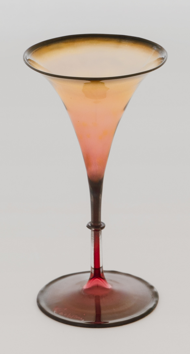 Likørglass i meget tynt, farget glass. Konisk trumpetformet kupa, hvor overgangen til den tynne stetten er markert med en ringformet vulst. Glasset hviler på en sirkulær fotplate. Likørglasset graderes i ulike fargenyanser: Foten, stetten og nedre del av kupa veksler mellom rosa og sort, sistnevnte med en metallisk glans. På øvre del av kupa blir glasset gul-oransje, og munningen er markert med sort.
