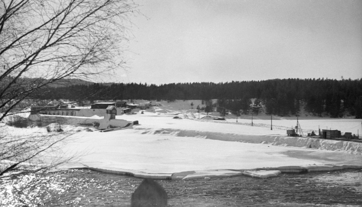 Anleggsarbeid på den midtre delen av den drøyt 300 meter lange terskeldammen ved Skjefstadfossen i Heradsbygda i Elverum, Hedmark. Fotografiet ble tatt i slutten av februar måned i 1942. Da var elva, med unntak av det østre løpet (i forgrunnen, der vannføringa var størst) is- og snødekt. Anleggsarbeiderne hadde rigget seg til med brakker ved den midtre delen av dammen, og de hadde fått strømforsyning og oppkjørt veg ut til arbeidsstedet. Sannsynligvis skulle de forsterke dammen, som på dette tidspunktet var drøyt 30 år gammel. Slikt arbeid ble gjerne gjort i vintesesongen, for da var vannføringa i elva liten. Til venstre i bildet, bak og under noen bladløse kvister, skimter vi sjølve kraftverksdammen og tilhørende bygninger ved vestre elvebredd.