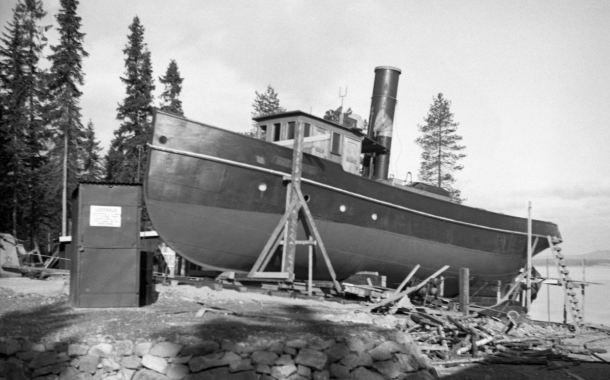 Tømmerslepebåten Trysilknut på slippen ved Sørlistøa høsten 1941.  Båten er trukket opp på land, ikke bare for vinterlagring, men etter stillasarbeidet å dømme også for skraping og maling.  I forgrunnen en natursteinsmur.  Til venstre for (like foran) baugen på båten et lite jernplatebeslått skur. Det omsluttet ei pumpe for petroleum.

Trysilknut ble bygd av Glommens mek. Verksted i Fredrikstad høsten 1913 og på nyåret i 1914. Navnet ble hentet fra en sagn, som nettopp i ble aktualisert ved at han ble hovedperson i folkelivsromanen «Trysil-Knut» av Rudolf Muus (1862-1935), som kom på markedet like før båten skulle sjøsettes.  Fortellinga ble gjenbrukt i manuskriptet til en film som kom året etter at dette fotografiet ble tatt, i 1942. Fartøyet fraktet komponentvis med jernbanen fra Fredrikstad til Rena og hentet derfra til Brevik ved Osensjøen med hester og sleder for sammenklinking.  Båten ble ombygd fra damp- til dieseldrift vinteren 1957-58.  



Motivet er tilnærmet identisk med SJF. 1990-01222