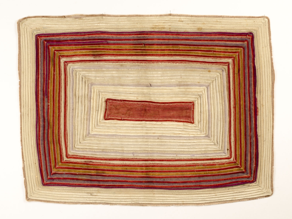 Matta sydd av fransband i beige, rött, blått, gult och lila. I mitten en rektangulär bit röd sammet. Baksida av kypertvävd kattun, röd med svart rosmönster.