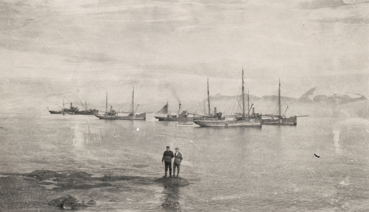 To menn står på land i Ny-Ålesund. I bakgrunnen er ei flåte med båter på fjorden.